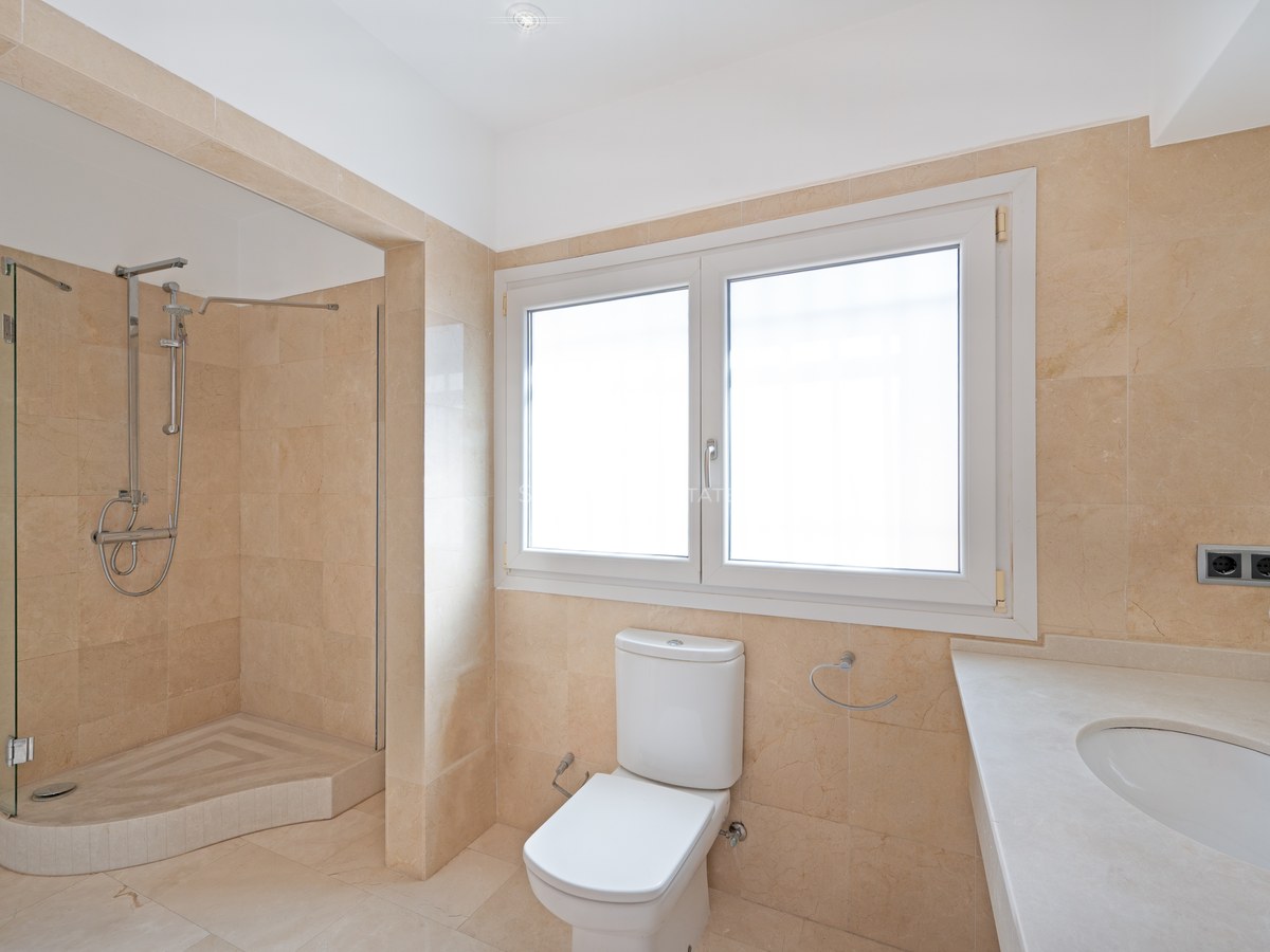 A modern bathroom featuring cream marble walls and floor, a glass walk-in shower, and a large frosted window.