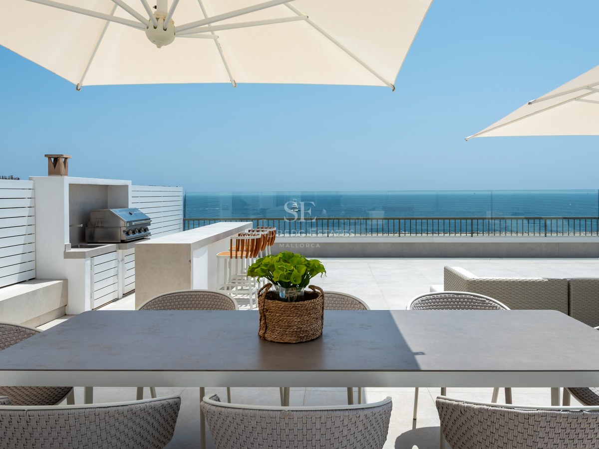 Luxury rooftop terrace featuring a dining area, outdoor kitchen, and glass railings overlooking the blue Mediterranean sea.