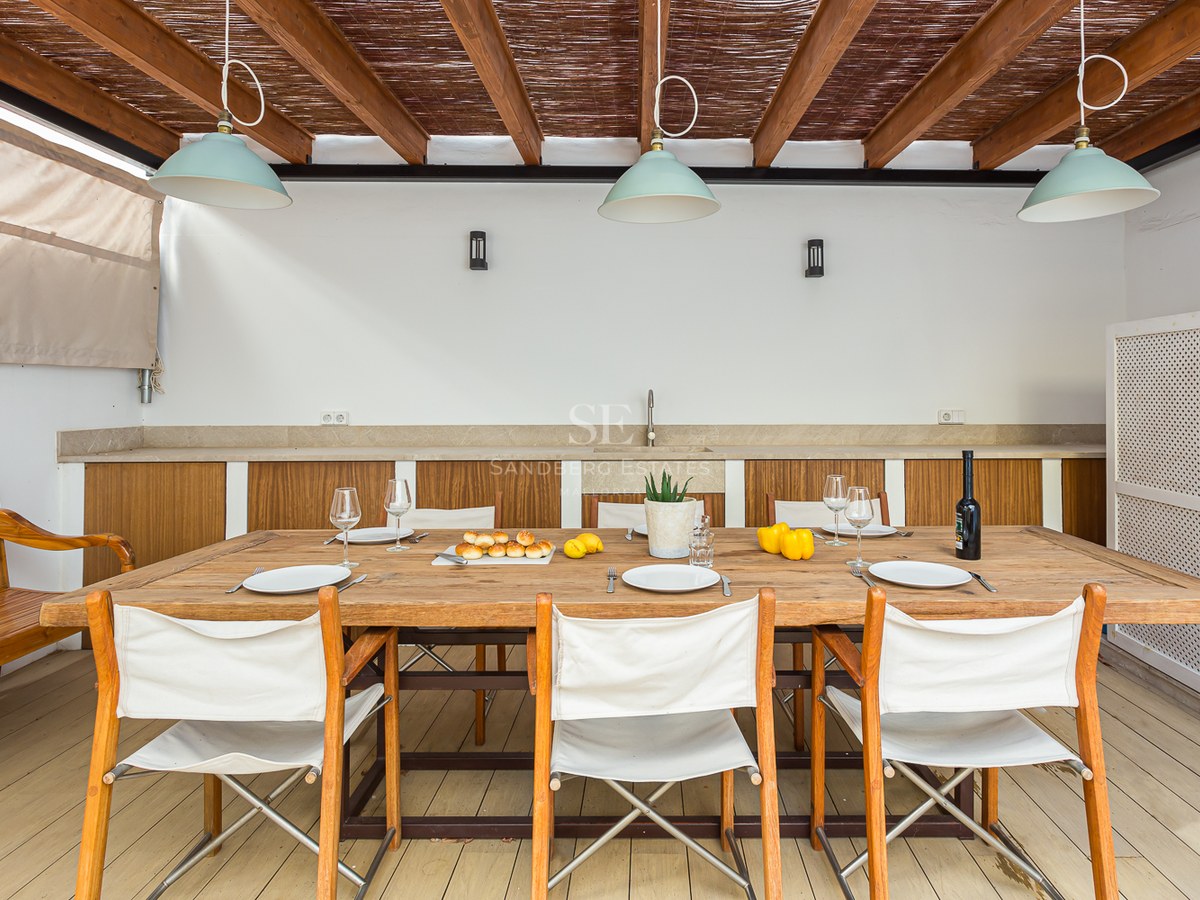 Terraza cubierta con gran mesa de madera, sillas de lona blancas y cocina exterior bajo una pérgola de madera.