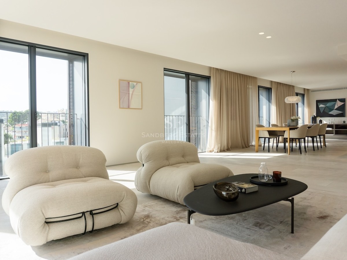 Ljust vardagsrum med designfåtöljer i beige, svart soffbord och matplats intill stora fönsterpartier.