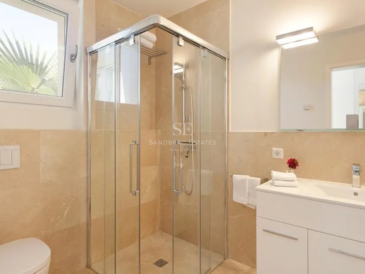 Bright bathroom featuring a glass corner shower, beige marble wall tiles, a white vanity unit, and a small window.
