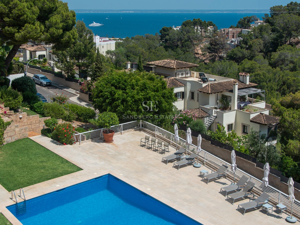 Flygvy över en blå pool och solterrass med solstolar med utsikt över Medelhavet och villor.