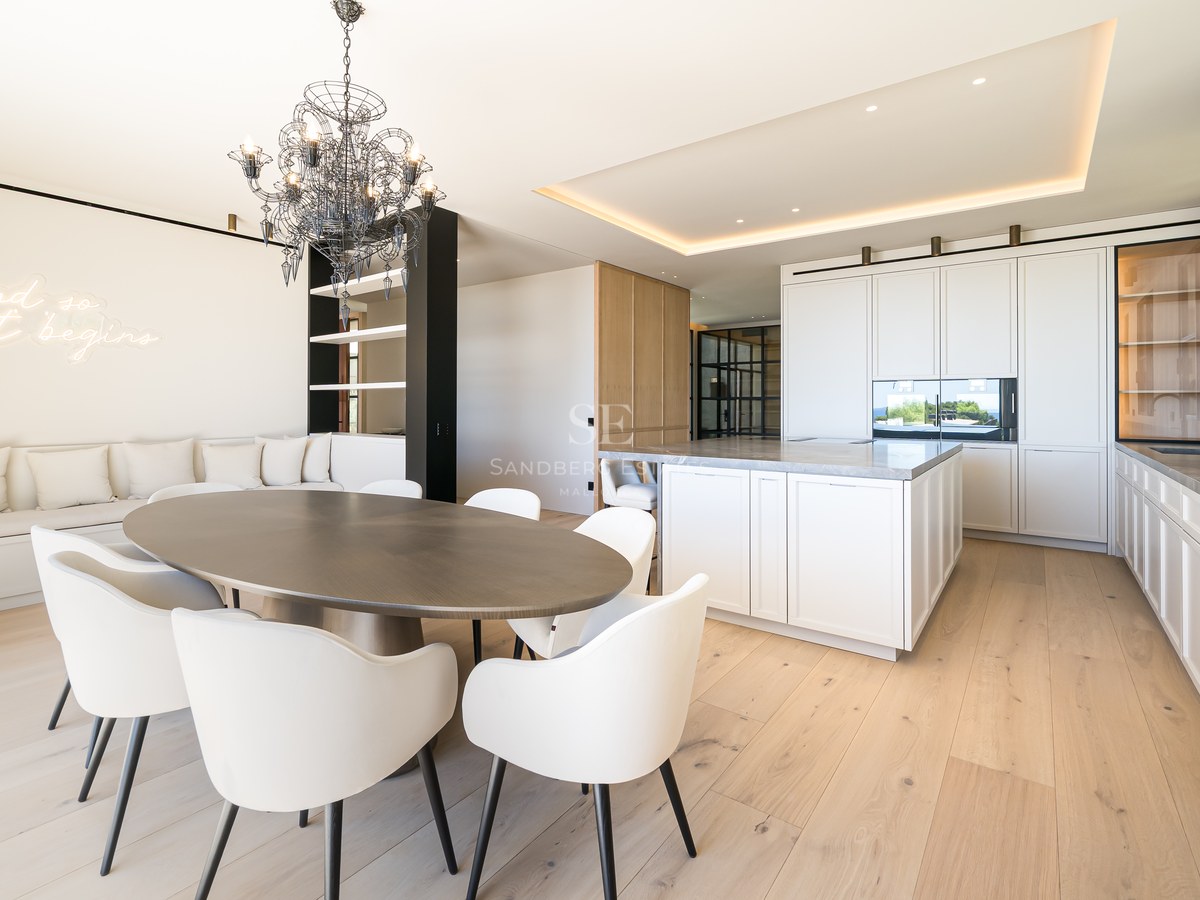 A spacious modern dining room featuring an oval wooden table, white chairs, and an integrated designer kitchen.