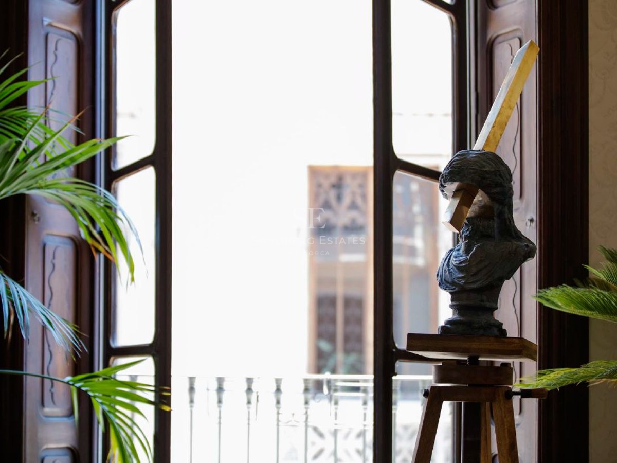 Sculpture of a bust on a wooden tripod stand next to a large bright window with green palm plants.