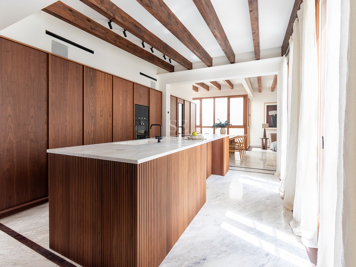 Modern kitchen with a fluted wood island, marble countertops, exposed ceiling beams, and natural light from large windows.