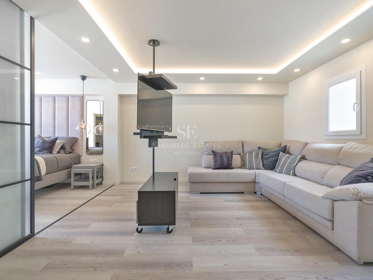Salon contemporain avec grand canapé en L beige, support TV pivotant et chambre adjacente séparée par une paroi vitrée.