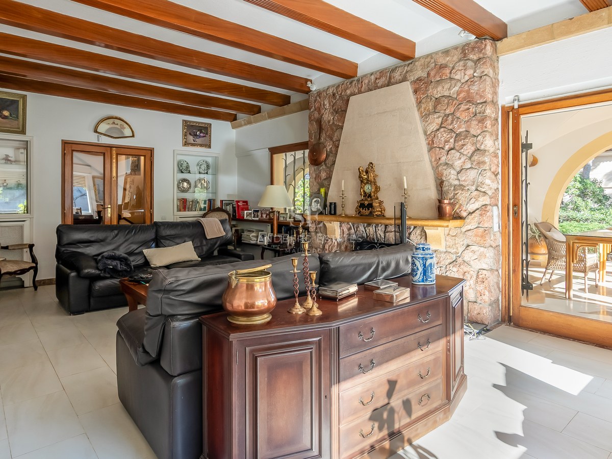 Acogedor salón con vigas de madera a la vista, gran chimenea de piedra, sofás de cuero negro y acceso a la terraza.