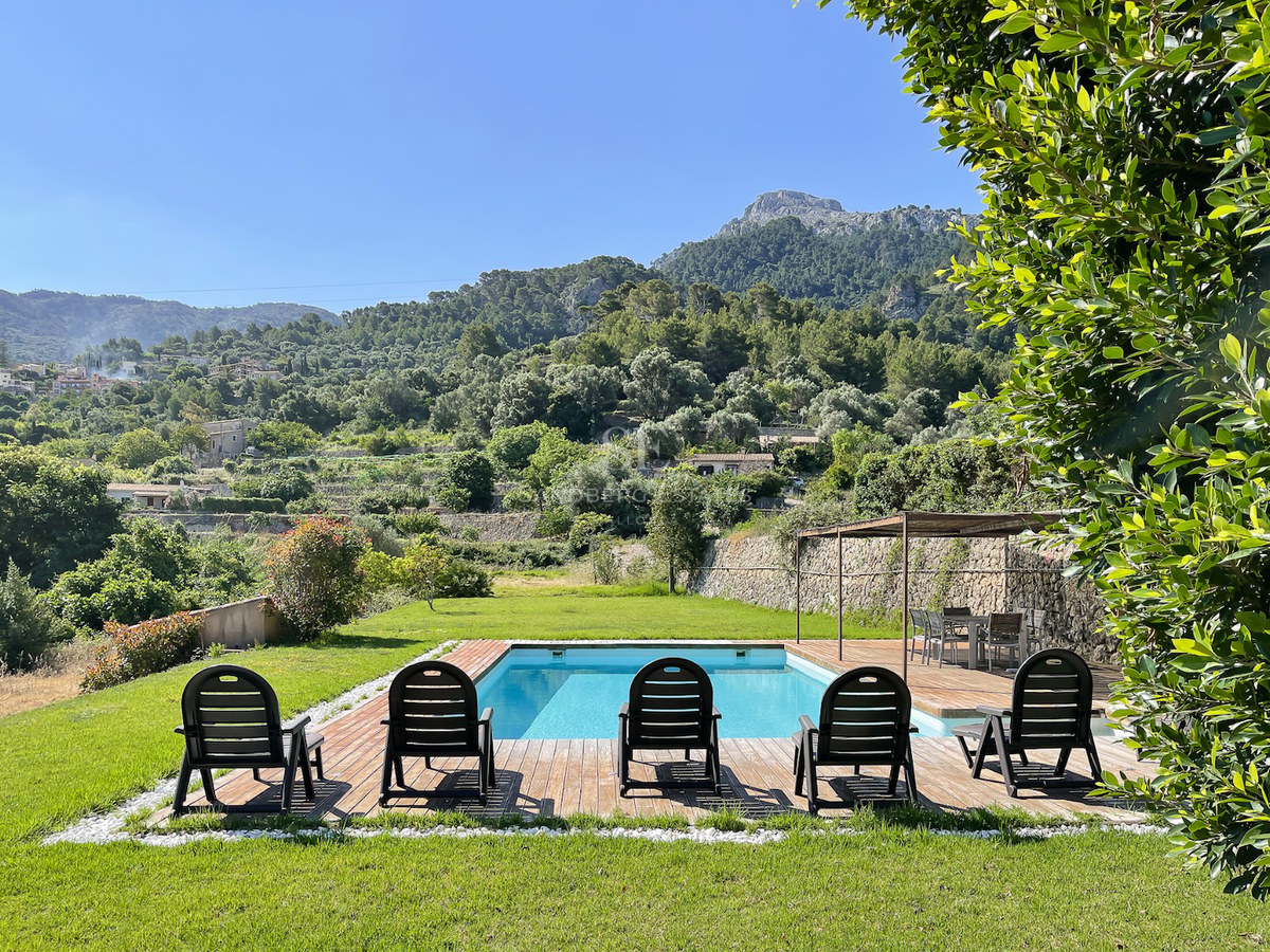 Piscina azul con terraza de madera y tumbonas frente a montañas verdes bajo un cielo despejado en Mallorca.