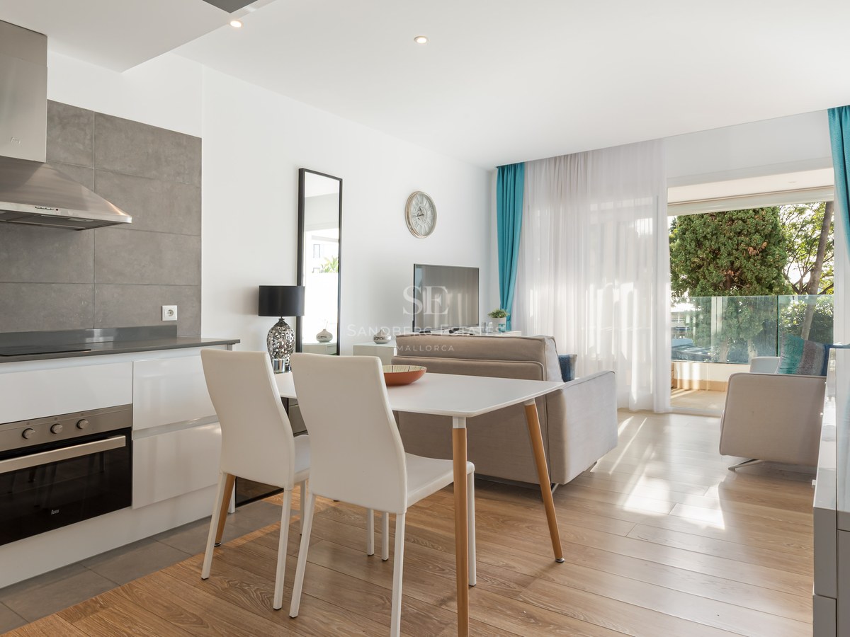 Bright open-plan room featuring a kitchen, dining area, and lounge leading to a sunny balcony with trees.