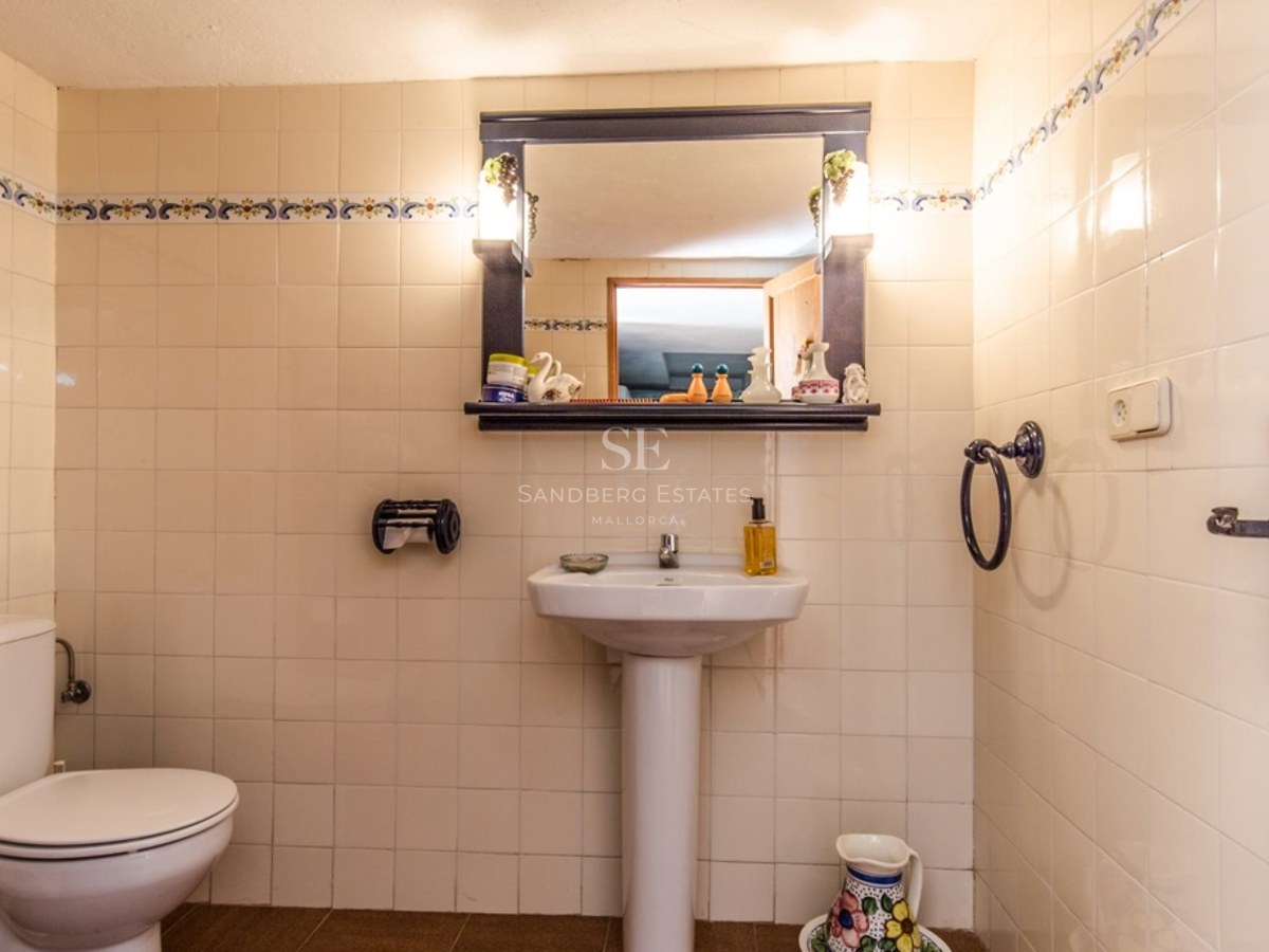 Salle de bain carrelée en crème avec lavabo sur colonne, toilettes et miroir à cadre sombre avec éclairage intégré.