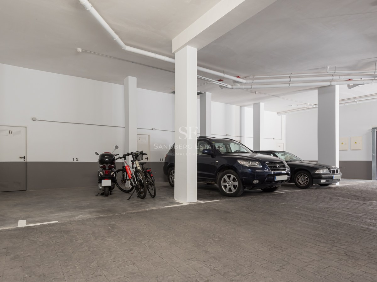 Underground parking garage with white pillars, grey textured stone flooring, and cars parked under artificial lighting.