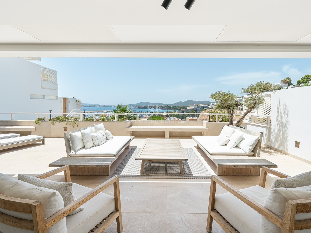 Grande terrasse blanche avec canapés et fauteuils en bois surplombant la mer bleue et la côte par une journée ensoleillée.