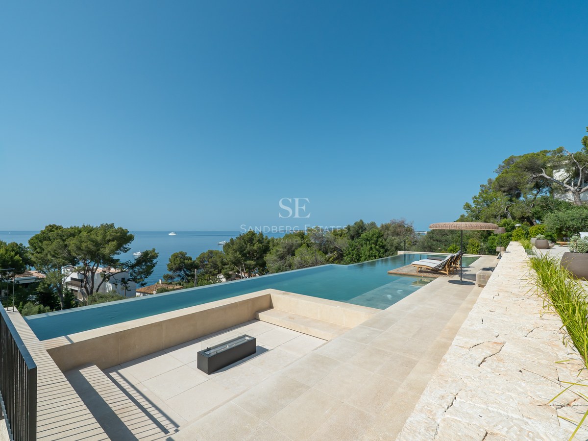 High-angle view of a luxury infinity pool and sunken fire pit area overlooking the Mediterranean sea and pine trees.
