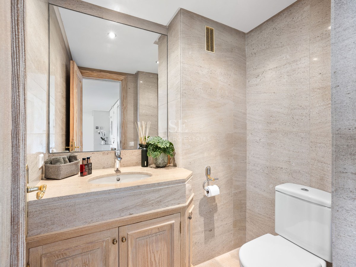 Salle de bains moderne avec carrelage en pierre beige, grand miroir et meuble sous vasque en bois.