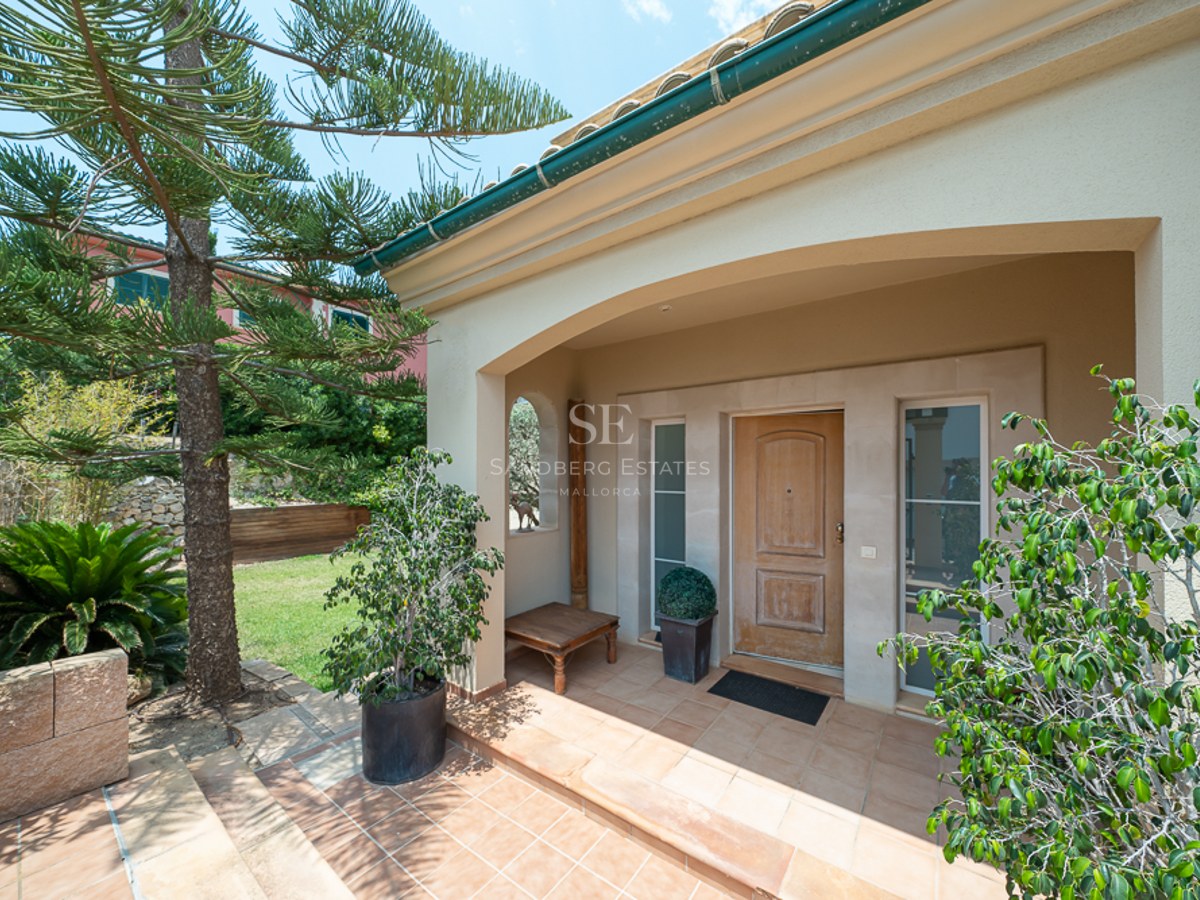 Entrée de villa voûtée avec porte en bois, sol en terre cuite et végétation luxuriante sous un ciel dégagé.