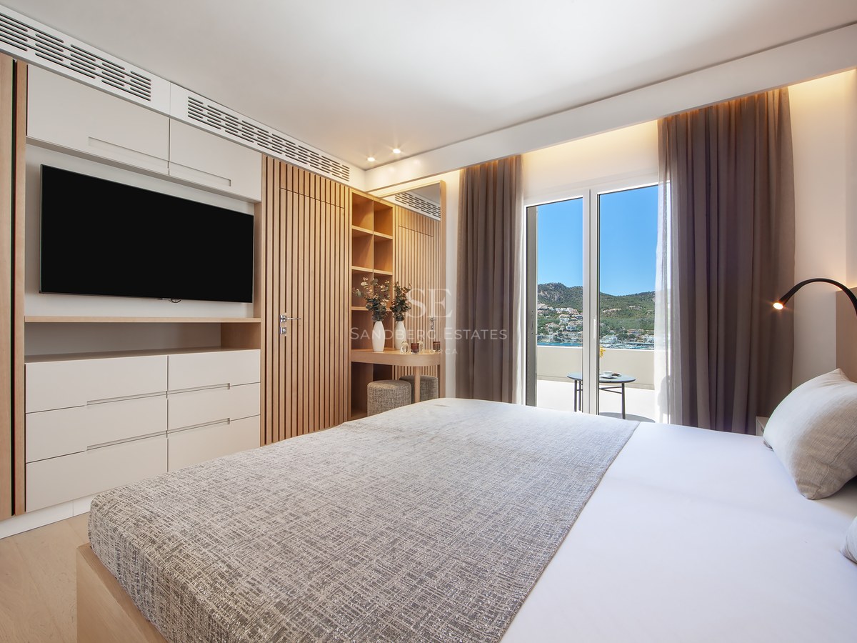 Bright bedroom with large bed, wood slat panels, wall-mounted TV, and balcony overlooking coast and mountains.