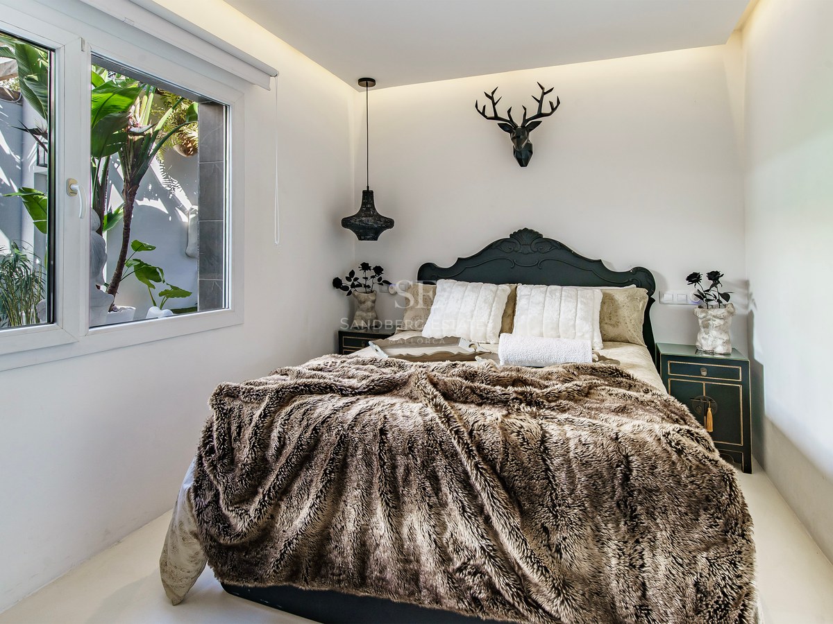 Chambre avec jeté de lit en fourrure, tête de lit sombre, tête de cerf décorative et fenêtre sur verdure.