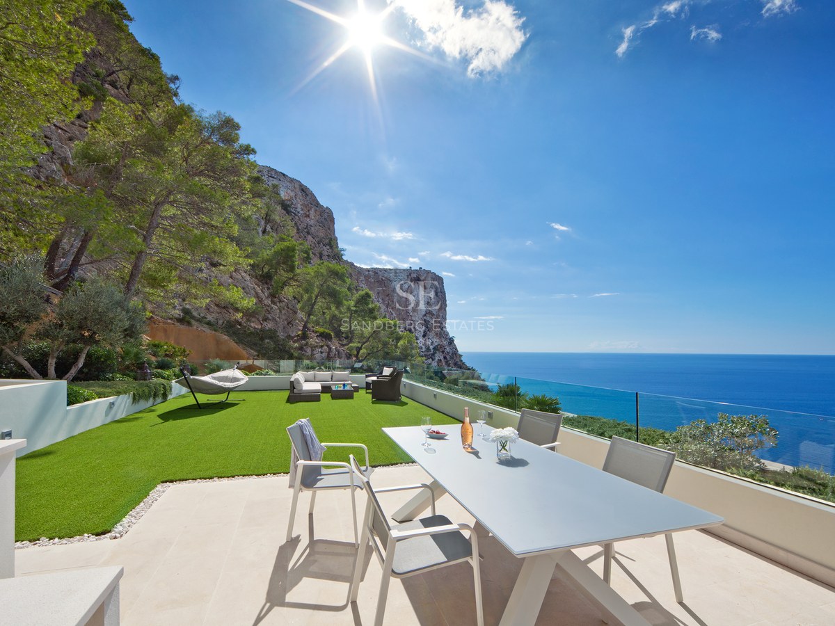 Modern outdoor terrace with dining area, artificial lawn, and glass railing overlooking the sea and cliffs.