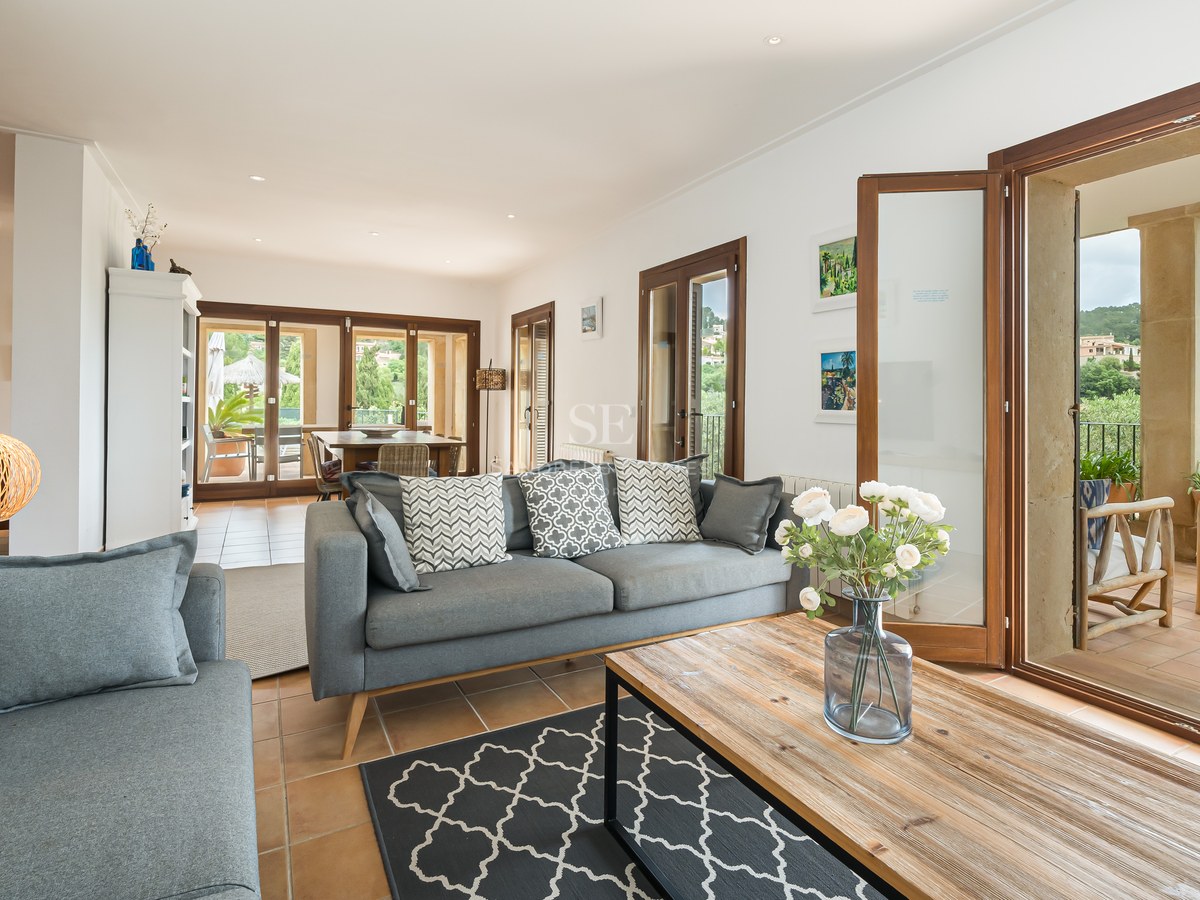 Modern grå soffa och soffbord i trä i ett ljust vardagsrum med terrakottagolv och stora glasdörrar.