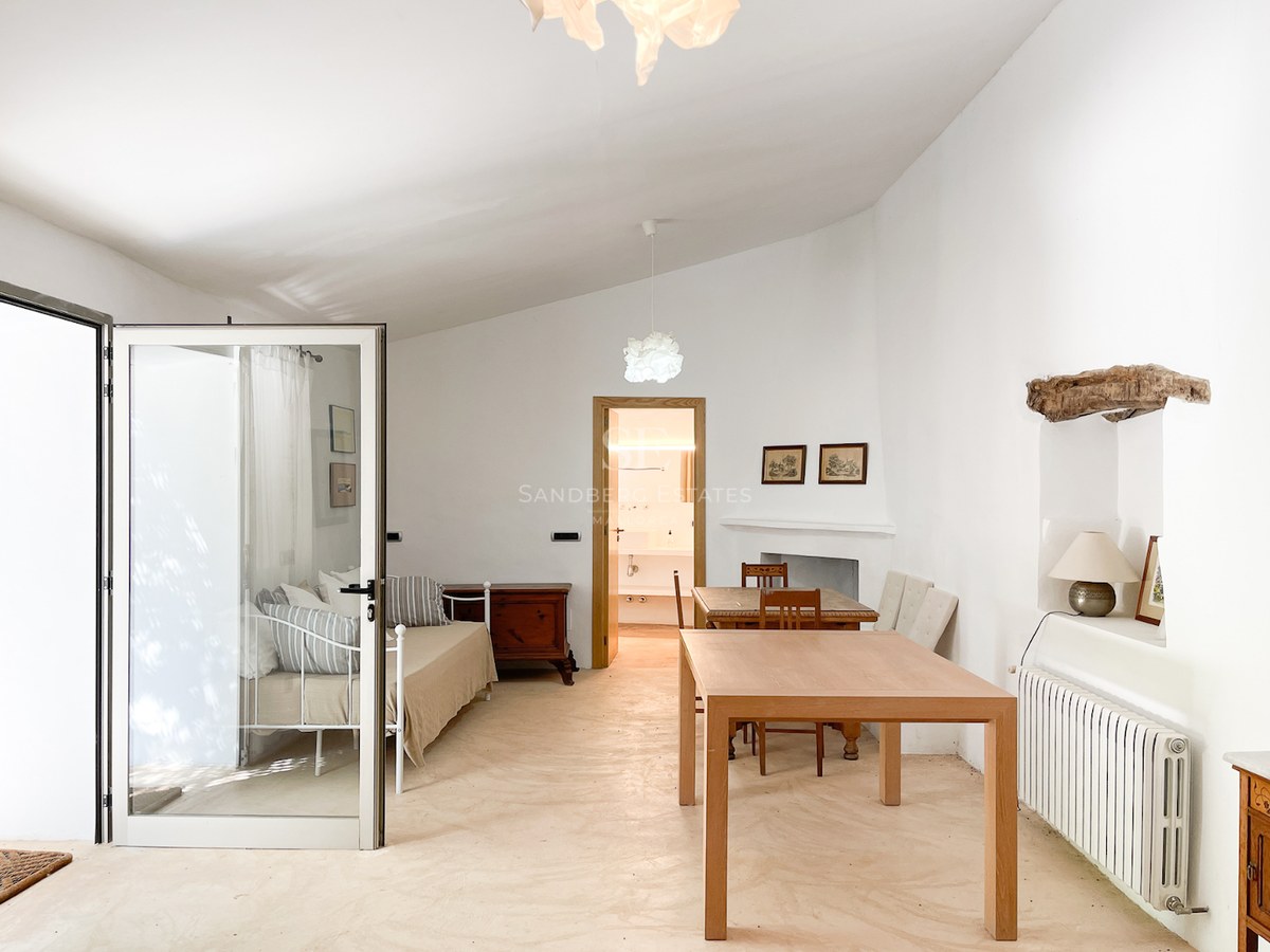 Dormitorio luminoso con cama de forja blanca, mesa de madera y suelos de microcemento bajo un techo abovedado.