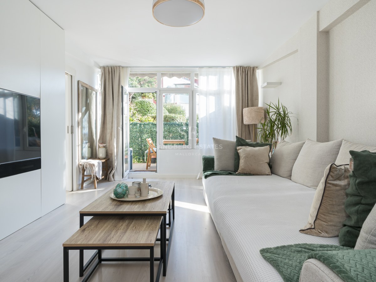 Bright living room featuring a large beige sofa, wooden coffee tables, and glass doors opening to a green terrace.