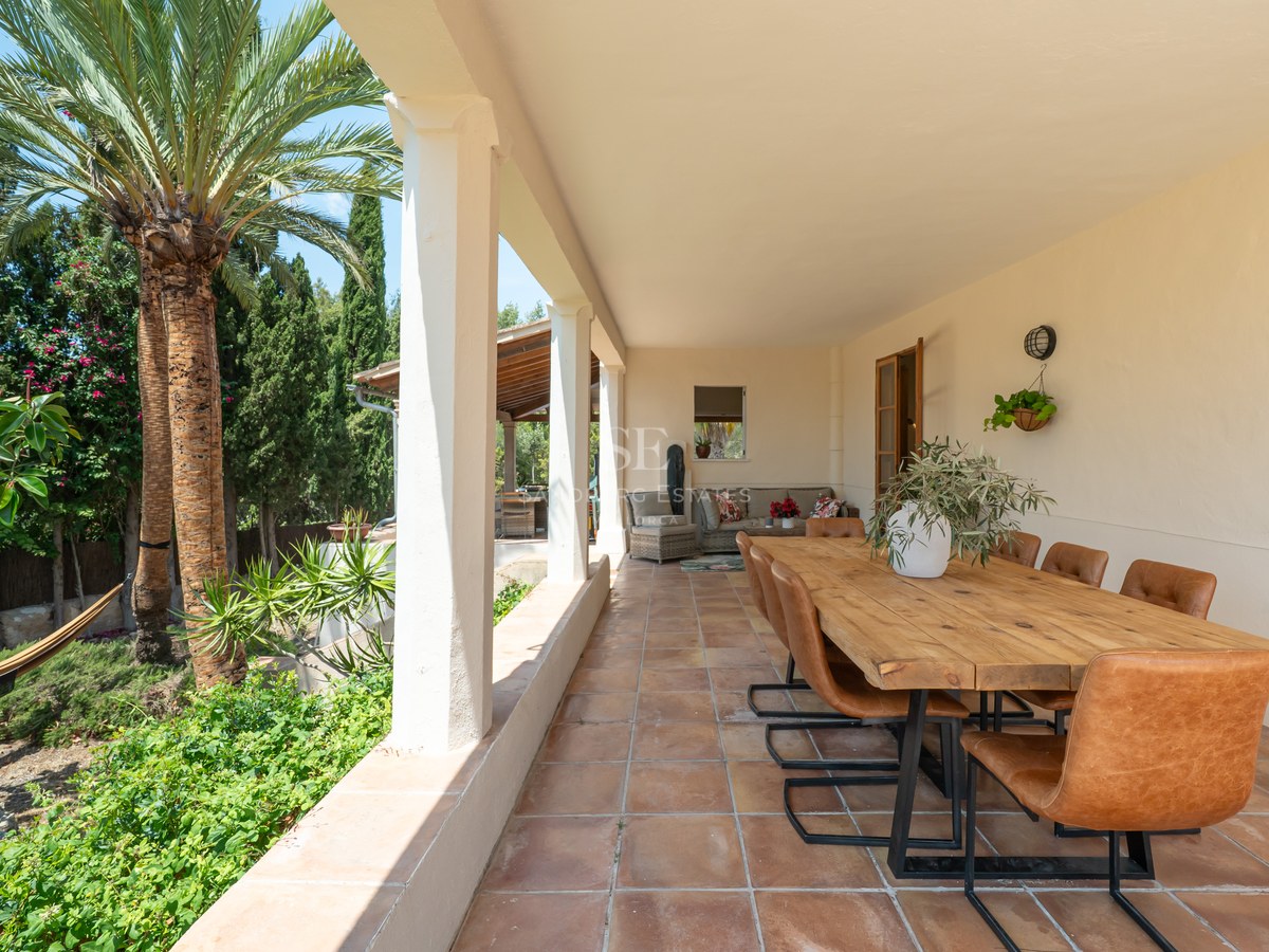 A covered terrace with a long wooden dining table, cognac leather chairs, terracotta floors, and lush garden views.
