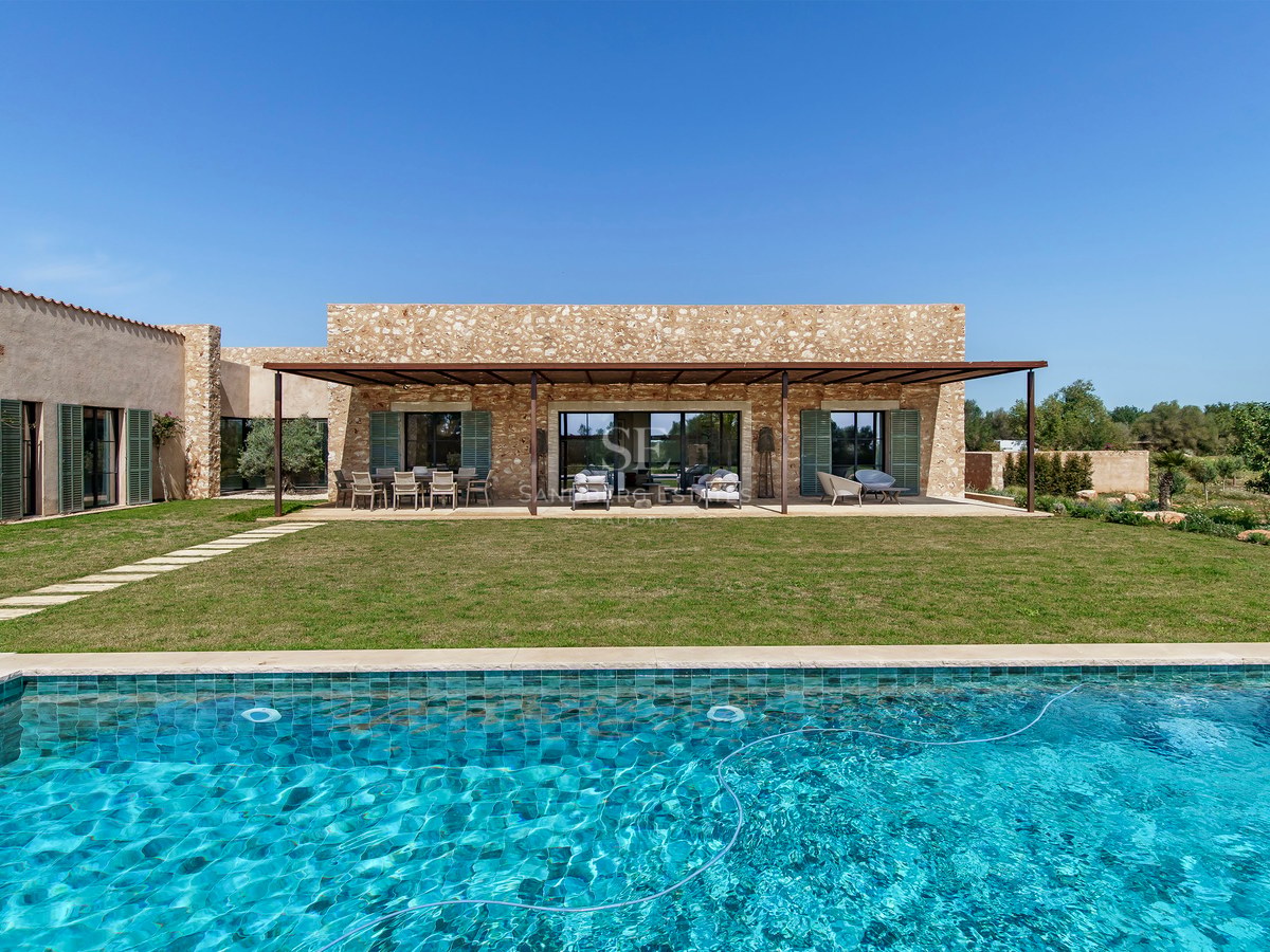 Extérieur d'une villa de luxe avec piscine turquoise, pelouse et façade en pierre naturelle sous un ciel bleu.