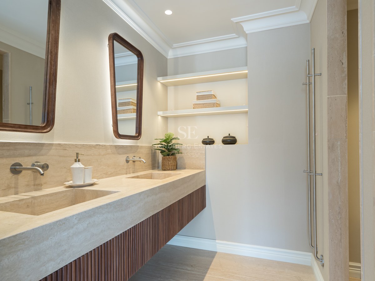 Baño moderno con lavabo doble de piedra travertino, armarios de madera y estanterías con iluminación LED.