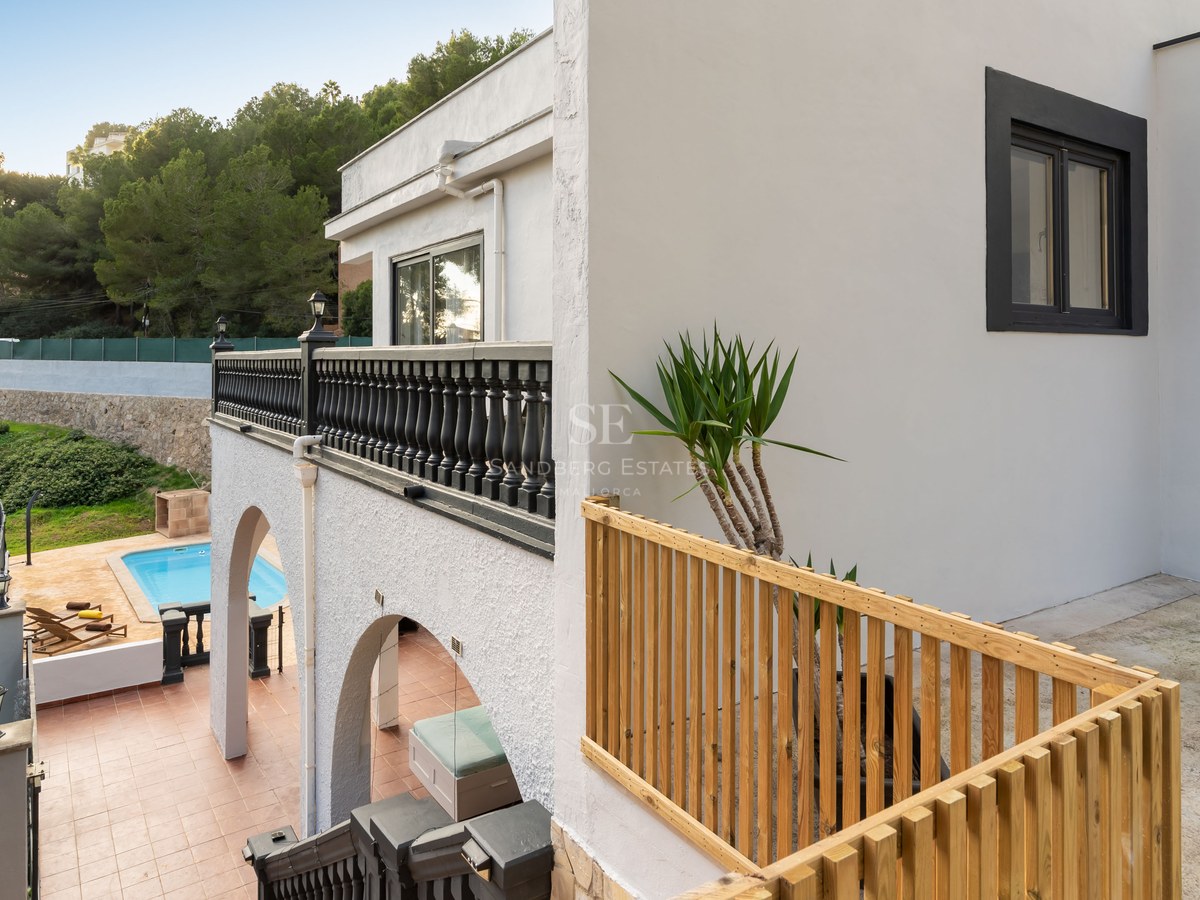 White multi-level villa with black and wooden railings, overlooking a turquoise pool and hillside forest.
