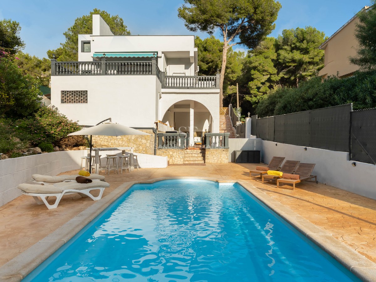 A blue rectangular swimming pool surrounded by a stone patio with sun loungers and a white villa in the background.