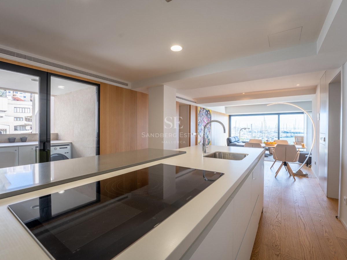 Îlot de cuisine blanc moderne avec plaque à induction et évier, donnant sur une salle à manger et vue sur le port.