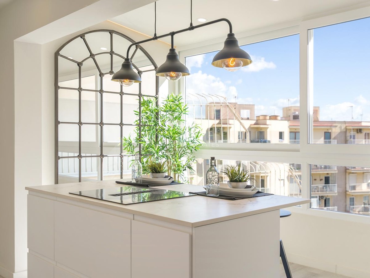 Îlot de cuisine blanc avec plaque à induction, suspensions industrielles et grandes fenêtres vue ville.