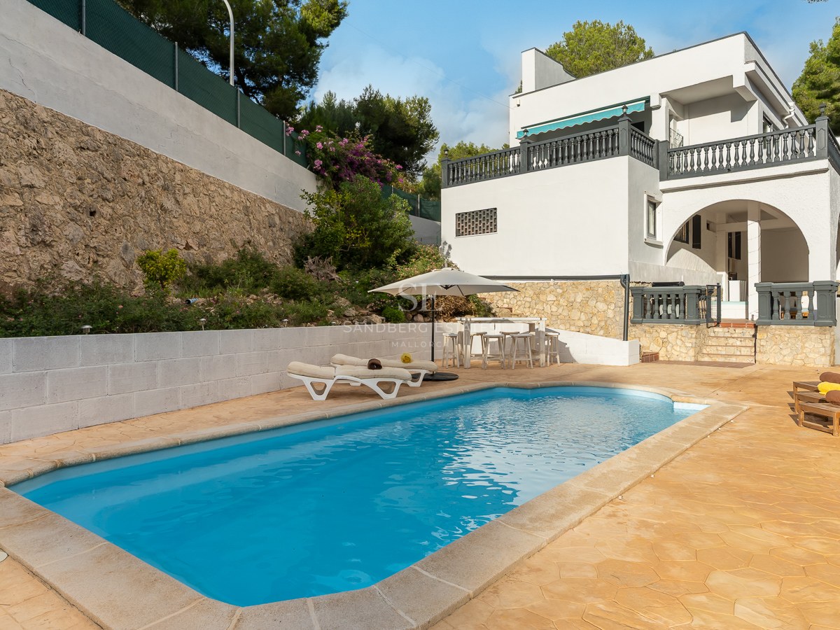 Rectangular swimming pool with turquoise water surrounded by a stone deck, sun loungers, and a white Mediterranean villa.