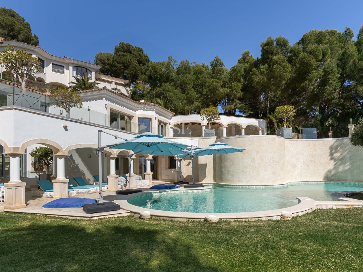 Curved swimming pool with turquoise umbrellas beside a white Mediterranean villa surrounded by pine trees.