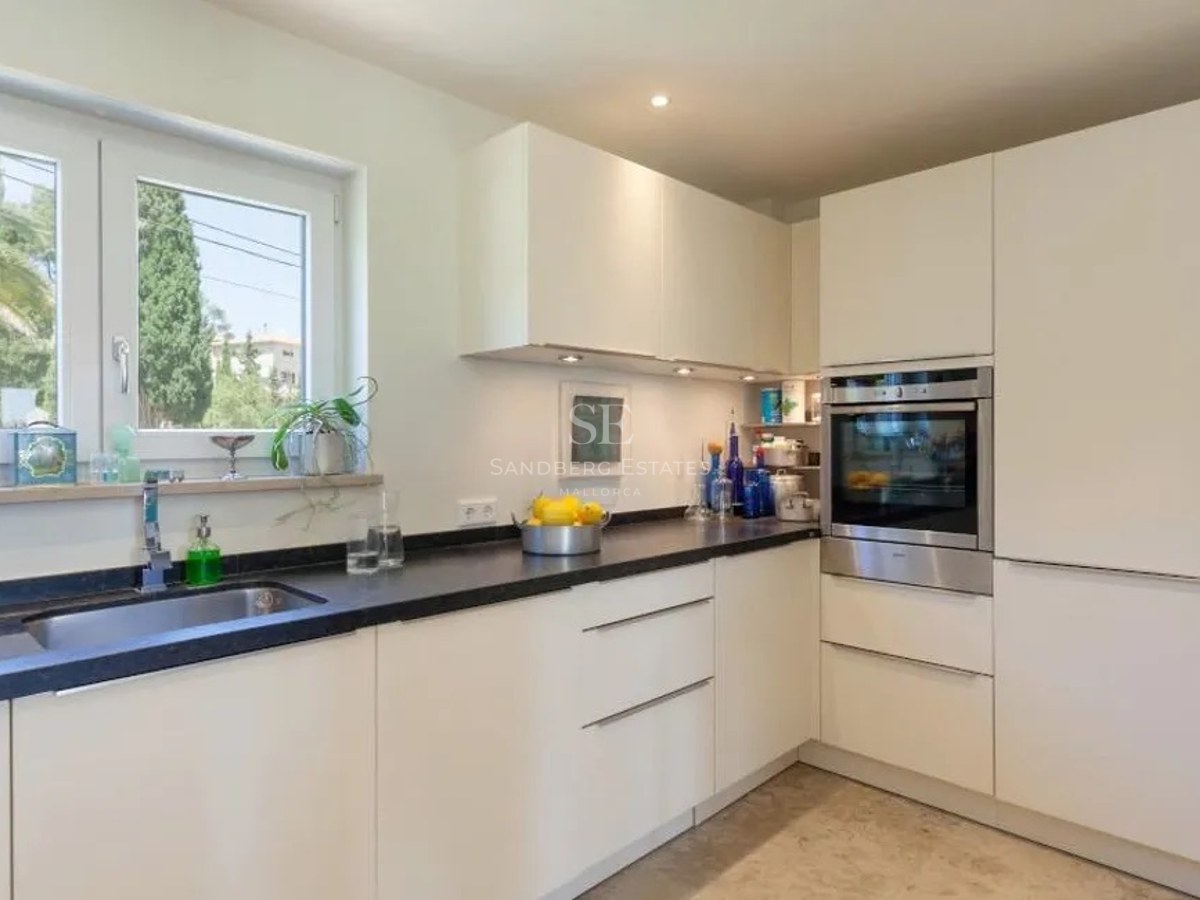 Contemporary kitchen featuring white handleless cabinetry, black granite countertops, and a window overlooking lush gardens.