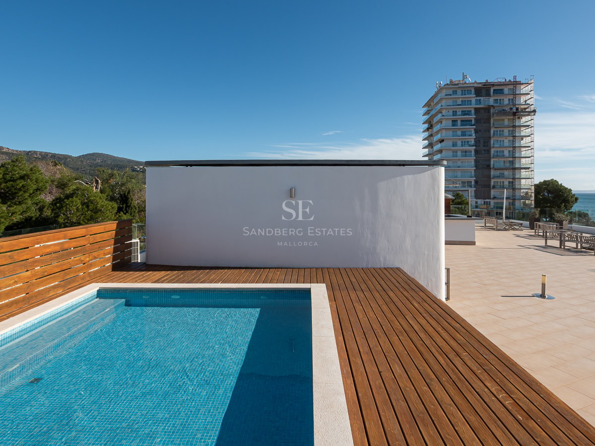 Modern rooftop swimming pool with wooden decking, overlooking the sea and mountains under a clear blue sky.