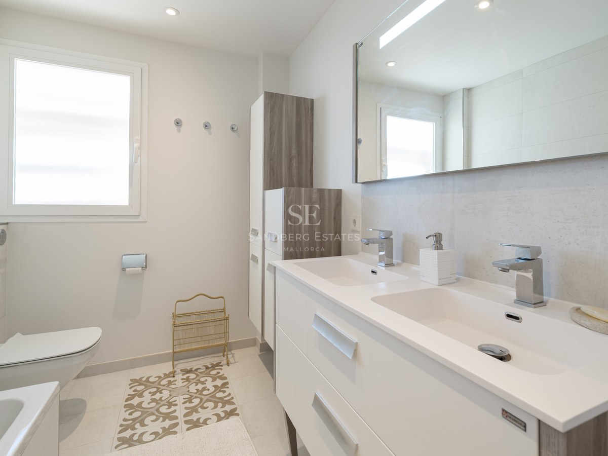 Salle de bain moderne lumineuse avec double vasque blanche, grand miroir, WC suspendu et lumière naturelle.