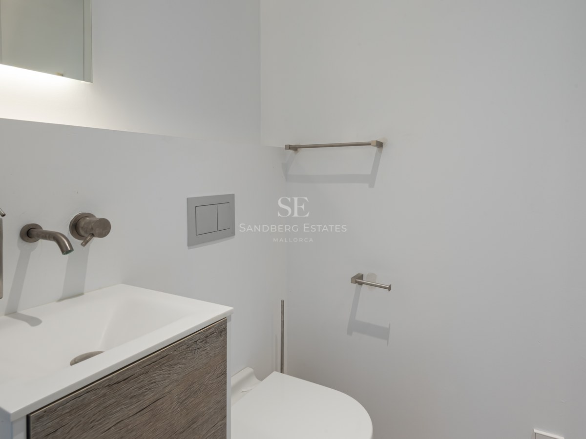 Detail of a white washbasin with brushed steel fixtures and a wooden vanity in a modern minimalist bathroom.