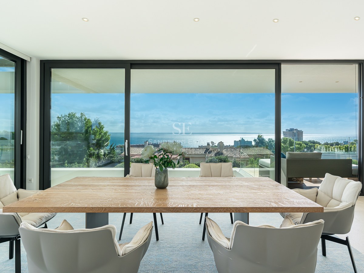 Salle à manger lumineuse avec table en bois et chaises grises face à des baies vitrées avec vue sur l'océan.