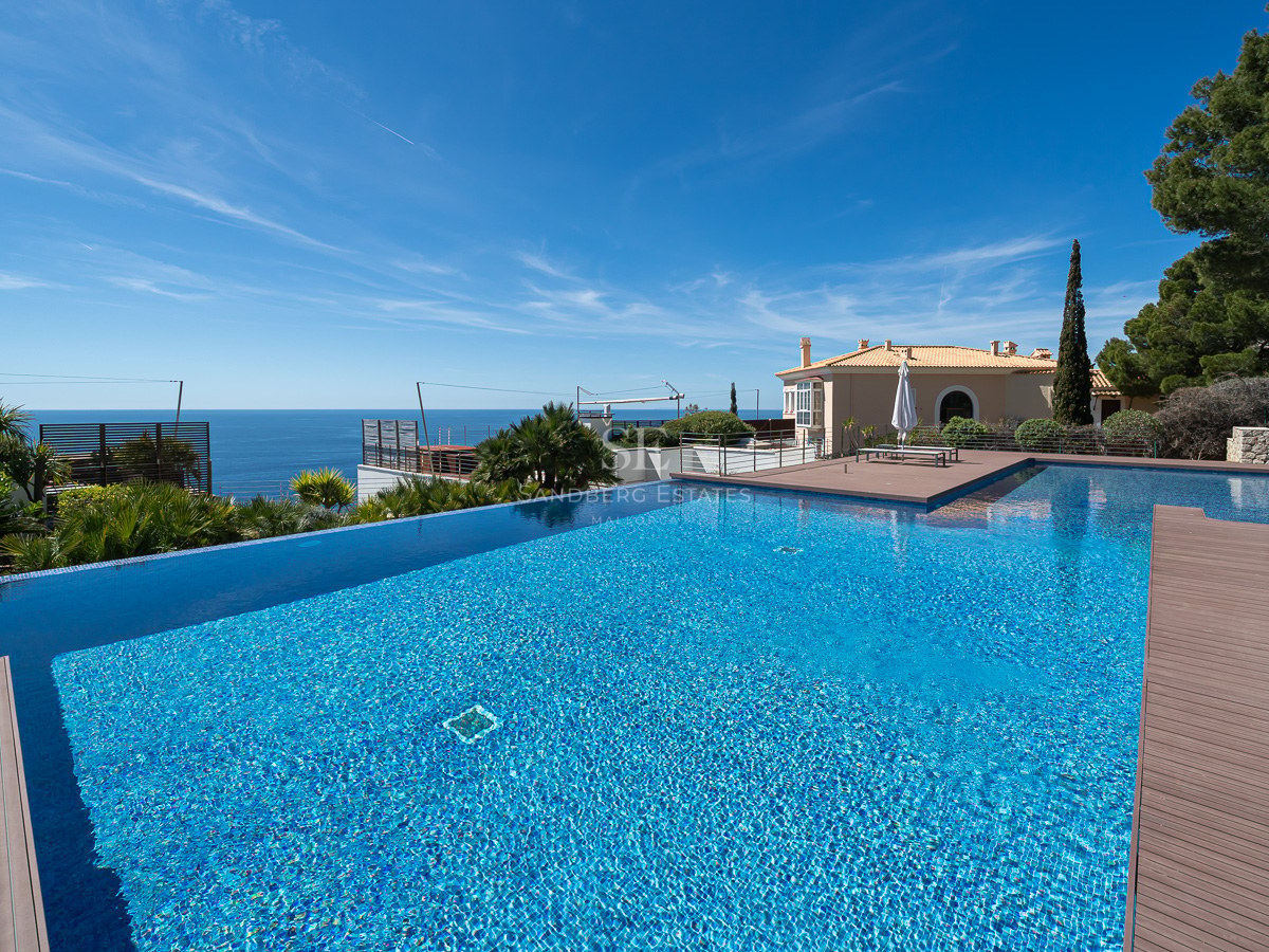 Large infinity swimming pool with blue tiles overlooking the sea and a stone terrace under a clear sky.