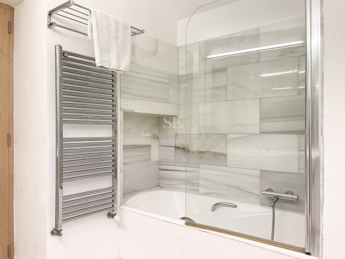 Bañera blanca con mampara de cristal, revestimiento de mármol gris y blanco y toallero radiador cromado.