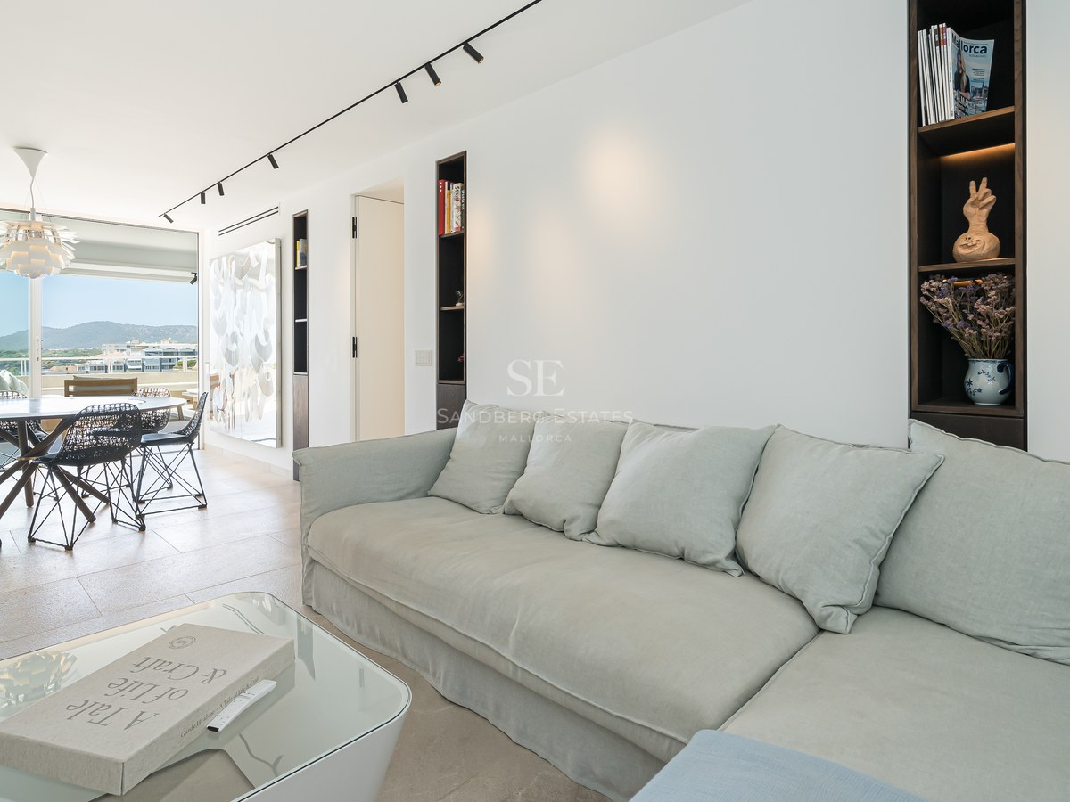 Salon lumineux avec un grand canapé gris clair, table basse en verre et salle à manger en arrière-plan avec vue sur la mer.