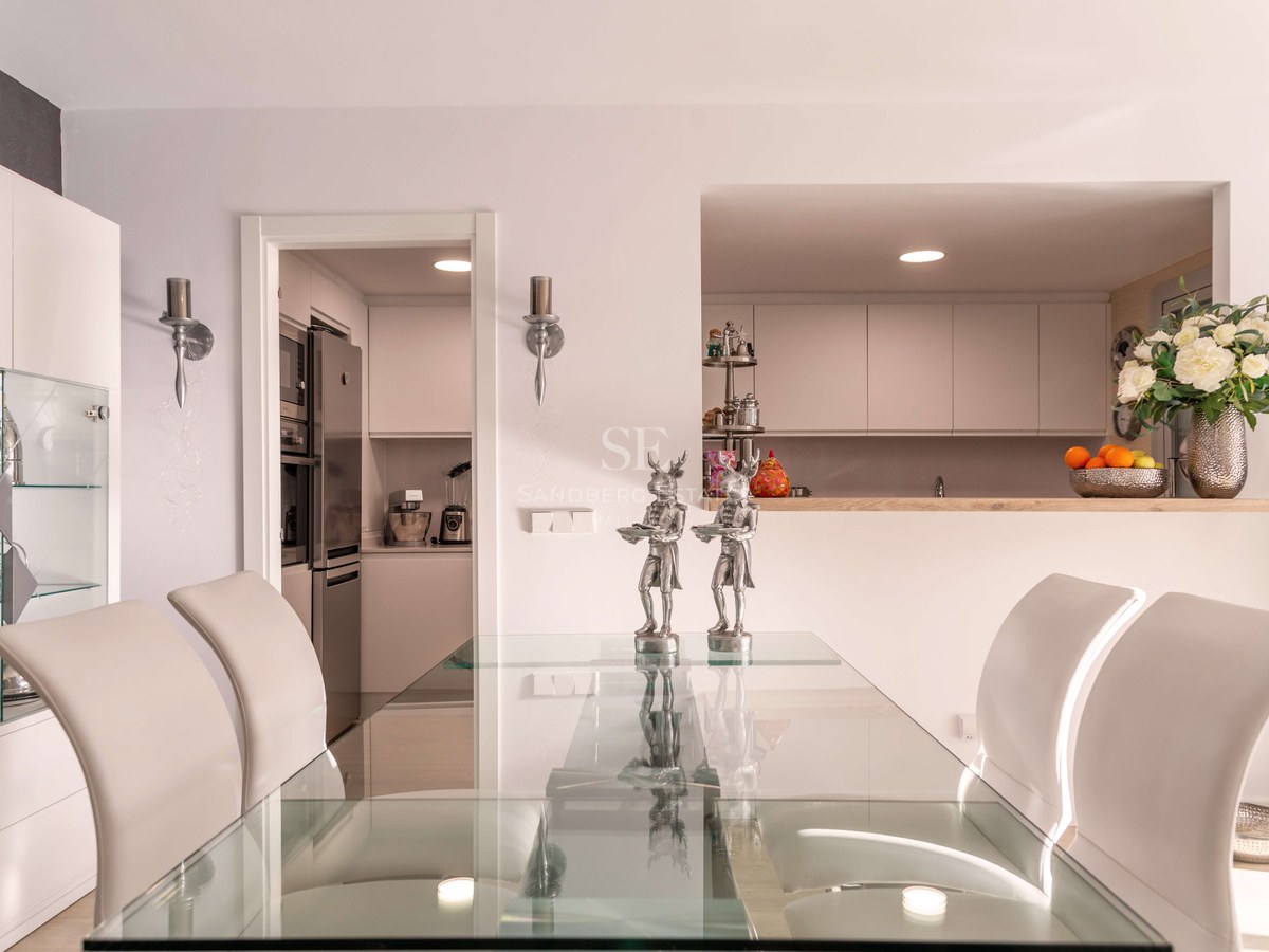 Salle à manger contemporaine avec table en verre, chaises blanches et vue sur une cuisine blanche moderne.