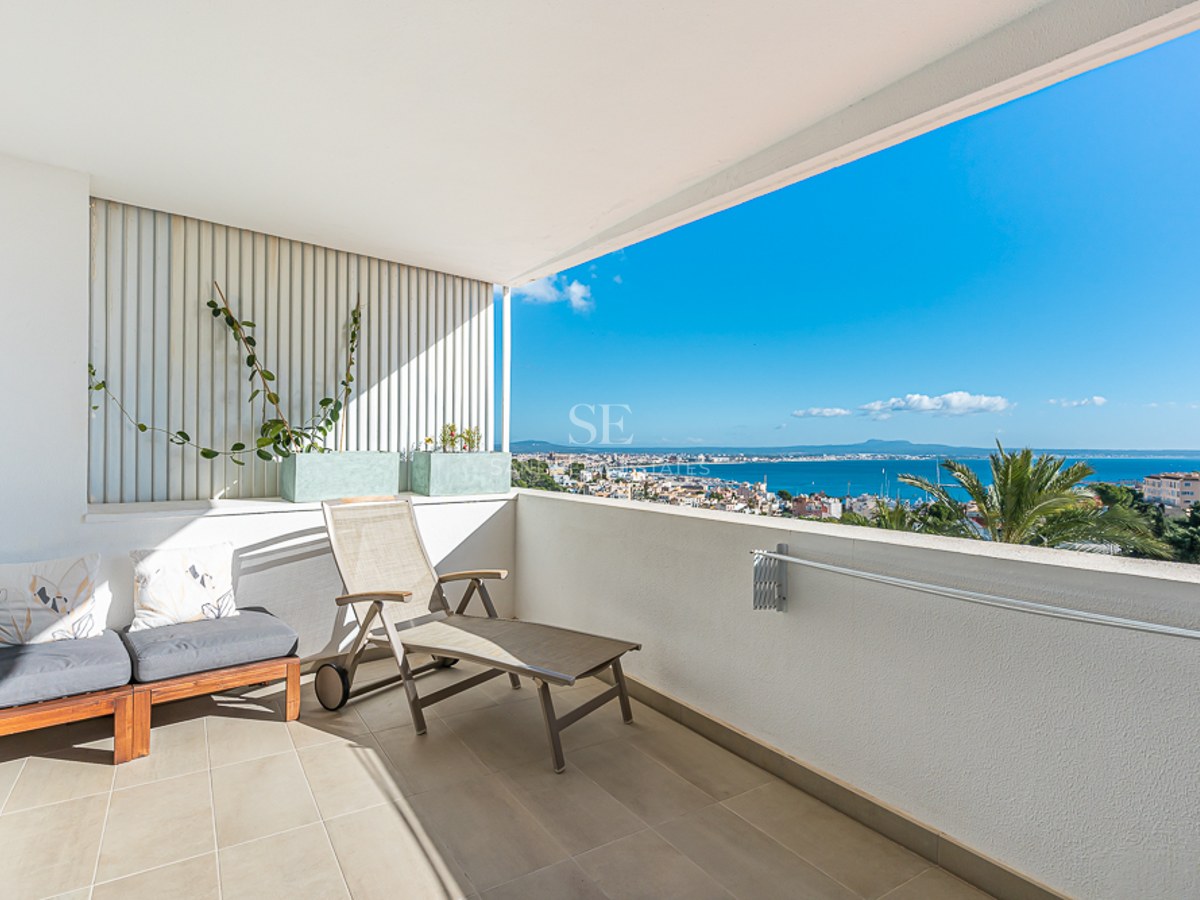 Spacious balcony with glass railing, panoramic sea views, and modern relaxation furniture. Plants add a natural touch.