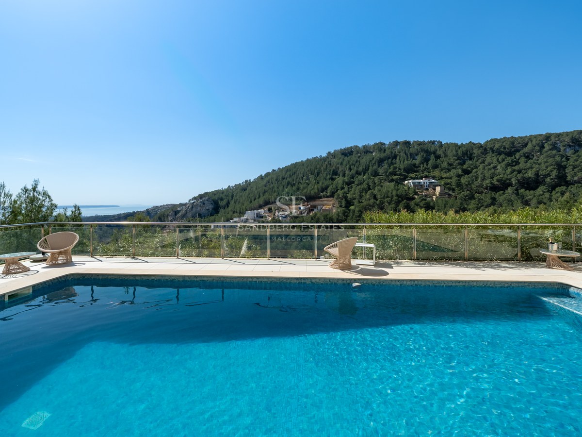 Piscina rectangular con terraza de piedra clara, barandilla de cristal y vistas a colinas boscosas bajo un cielo azul.