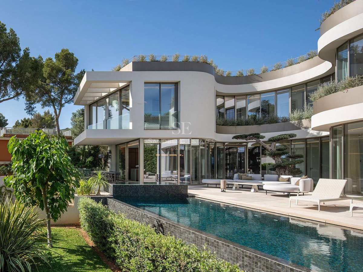 Modern vit villa med svängda väggar, en turkos infinitypool och utomhuslounge under en klarblå himmel.