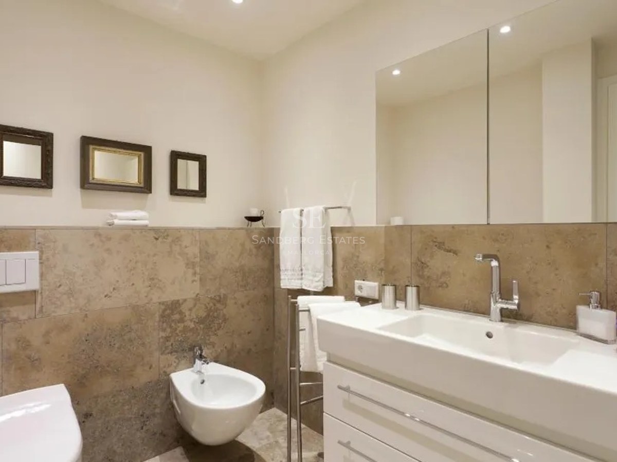 Modern bathroom featuring beige stone wall tiles, a white vanity unit, bidet, and a large wall-mounted mirror.