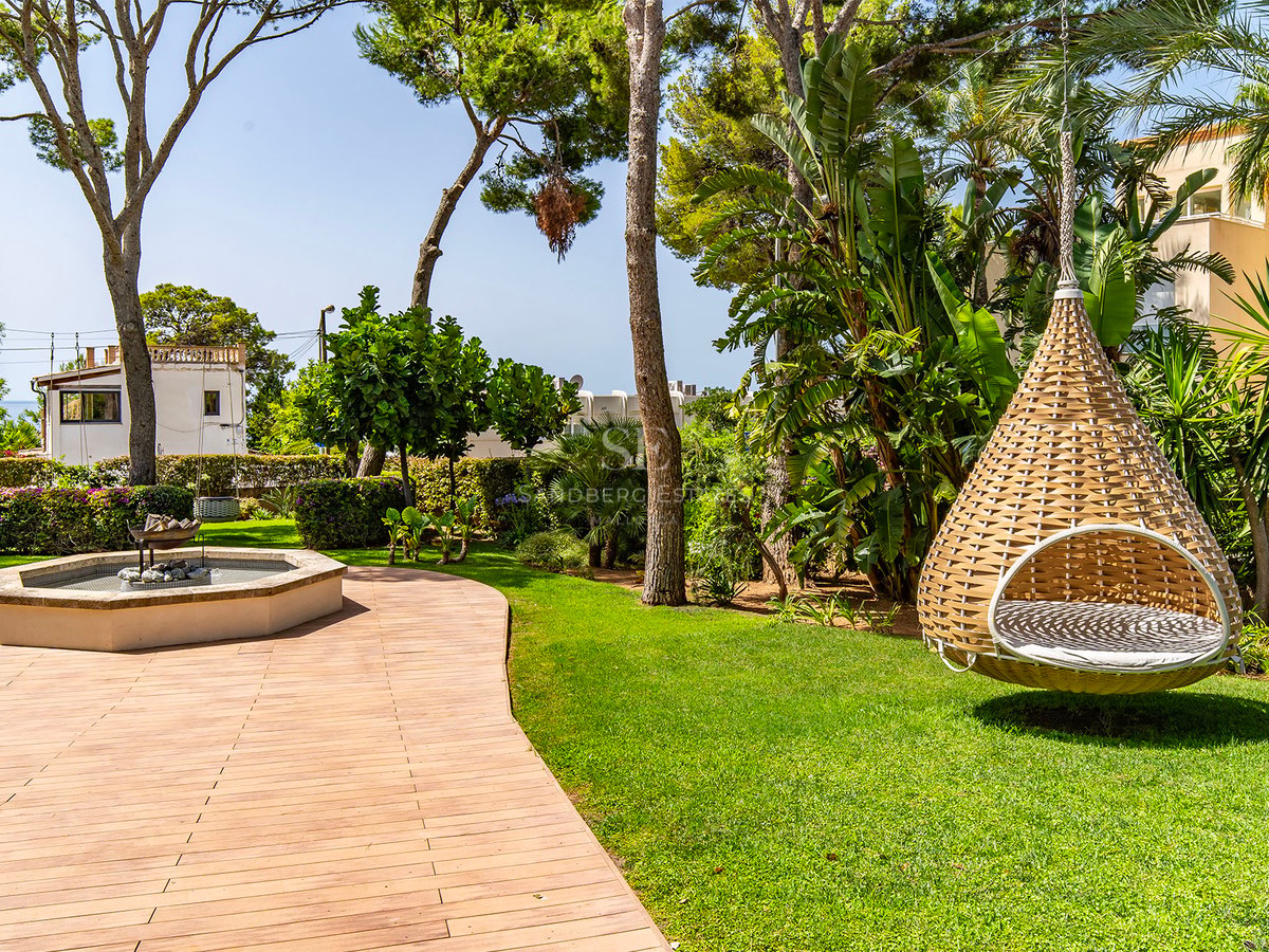 Jardin luxuriant avec terrasse en bois, pins parasols et fauteuil suspendu design sur une pelouse entretenue.