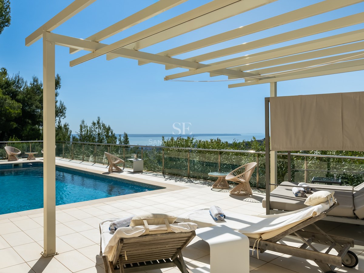 Terraza soleada con piscina, pérgola blanca, tumbonas y barandillas de cristal con vistas al Mediterráneo.