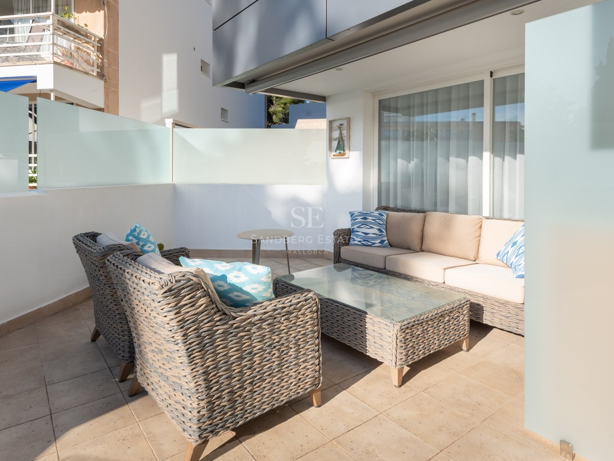 Modern terrace with grey wicker sofas, beige cushions, and blue pillows on a sunlit tiled patio with glass privacy walls.